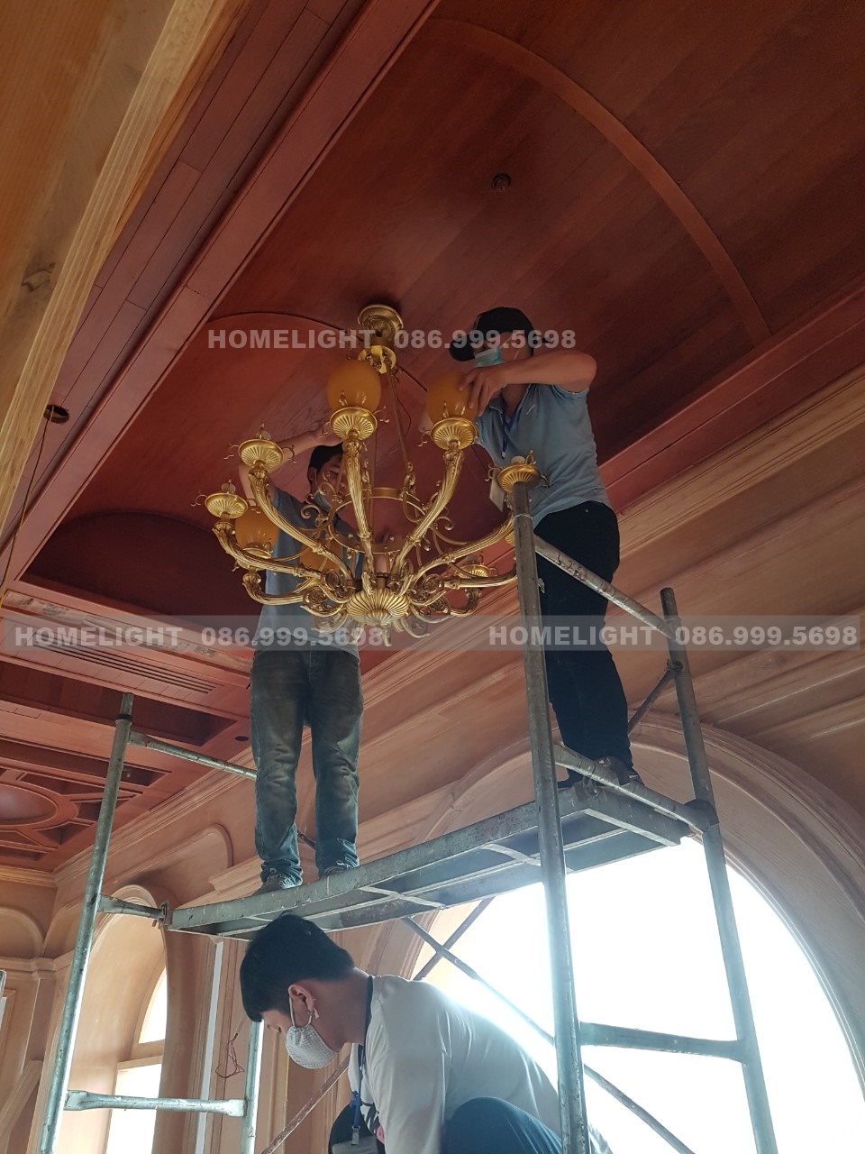 Thợ HomeLight trên giàn giáo lắp chao thuỷ tinh cho đèn chùm đồng dưới trần gỗ vòm, một bước thi công đèn trang trí tại sân golf Long Biên